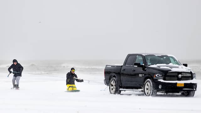 Twee jongens snowboarden achter een auto op het strand van Egmond.